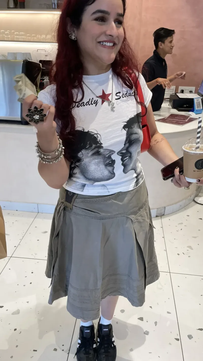 A guest at a cafe edition in a thrifted graphic tee and cargo skirt, iced coffee in hand.